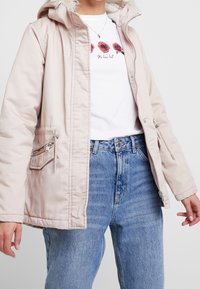 Ljusrosa huvjacka med fuskpälsdetaljer, zip-fickor och sydda detaljer. Bar över en vit t-shirt och blå jeans.