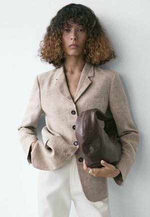 Donna con capelli ricci ombré che indossa un blazer beige e pantaloni bianchi, tiene in mano una borsa di pelle marrone scuro morbida su uno sfondo chiaro.