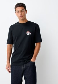 Črna bombažna majica s kratkimi rokavi, ki ima majhno grafično zasnovo risanega obraza s rdečo božično kapo na levem prsnem delu.