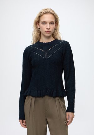 Femme aux cheveux blonds ondulés portant un pull en tricot noir avec un ourlet à volants et un pantalon plissé marron sur un fond uni.