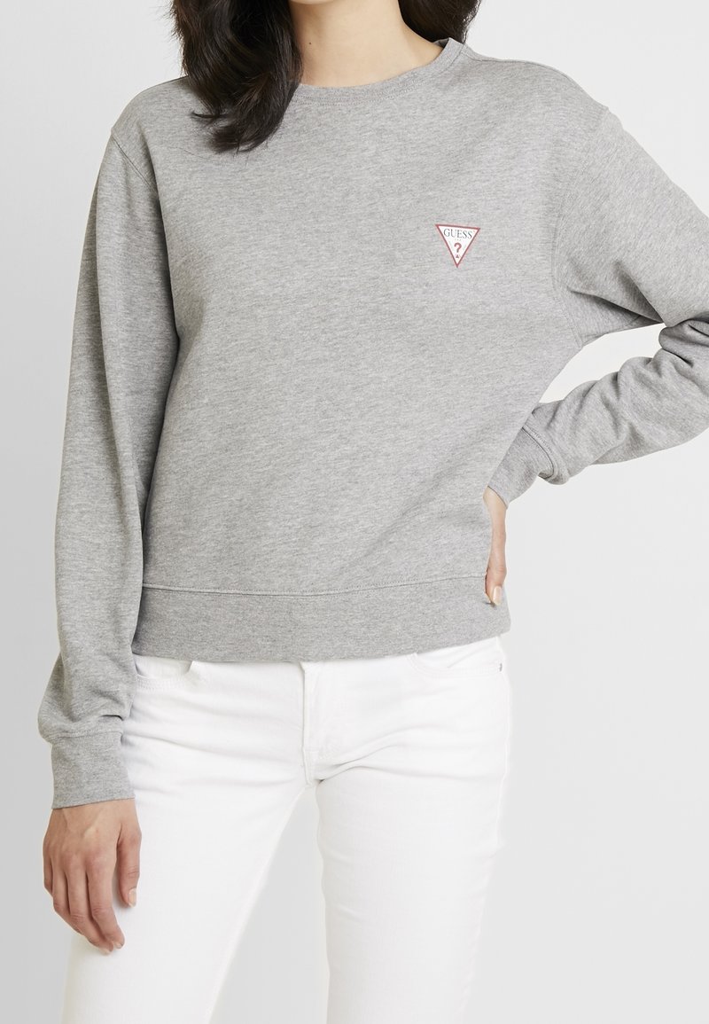 Femme portant un sweat Guess gris clair avec logo sur la poitrine et un pantalon blanc, posant avec une main sur la hanche sur fond uni.