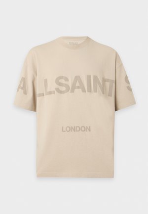 Un mannequin masculin porte une casquette marron, un tee-shirt beige avec un logo, un pantalon ample et des baskets marron sur un fond blanc uni.