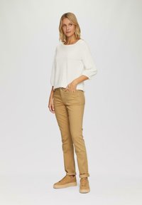 Weißes, langärmeliges, lockeres Hemd kombiniert mit beigen, schmal geschnittenen Hosen. Das Model trägt beige Sneaker. Weiche Textur, neutrale Farbpalette.