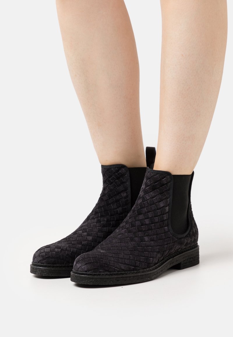 Jambes portant des bottines noires à texture tissée, avec des panneaux latéraux élastiques et des talons plats bas sur fond blanc.