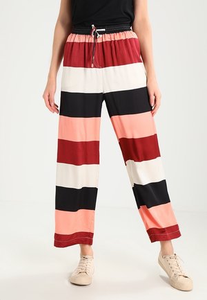 Kobieta ubrana w szerokie spodnie w poziome paski w kolorach czarnym, białym, czerwonym i brzoskwiniowym, zestawione z beżowymi trampkami i czarną bluzką.