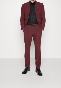 Bordowy garnitur z dopasowaną marynarką i spodniami ze ściągaczem, zestawiony z czarną koszulą. Całość dopełniają błyszczące czarne buty.