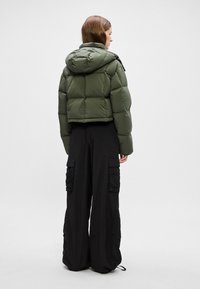La doudoune verte présente un design court, des sections matelassées horizontales et une capuche. Elle est complétée par un pantalon cargo noir à larges jambes et avec poches.