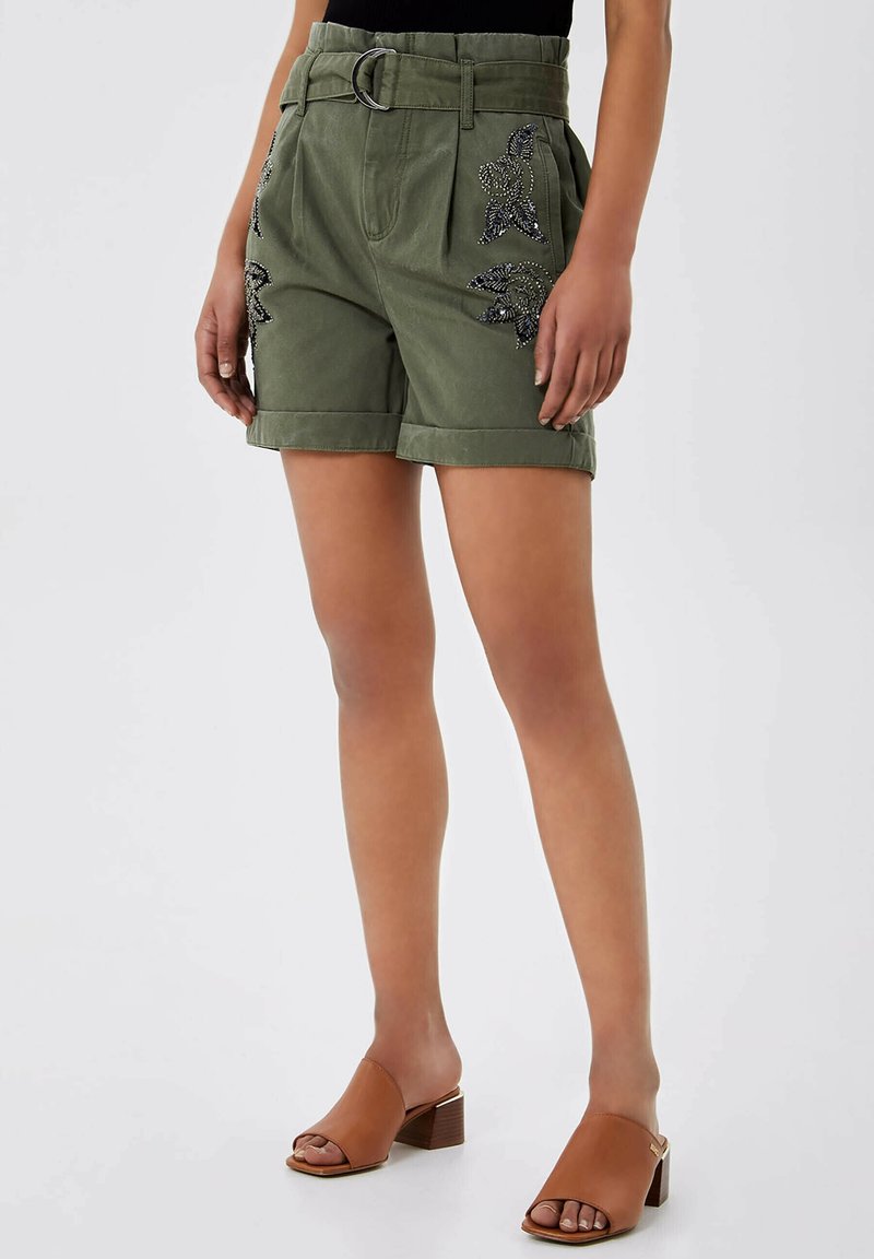 Frau trägt olivgrüne Shorts mit Gürtel und schwarzer Perlenblumenstickerei, kombiniert mit braunen Sandalen mit offenem Zehenbereich und Blockabsatz, stehend mit entspannten Händen.