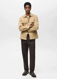 Khaki button-up skjorte med to brystlommer, lys tan tekstur; parret med mørkebrune bukser og sorte sko. Enkel, skræddersyet design.