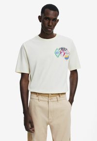 Short-sleeve white cotton t-shirt featuring colorful embroidered patches, round neckline, and a relaxed fit. Paired with beige pants.