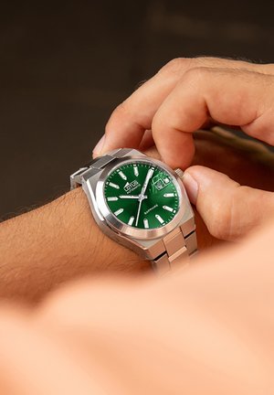 Montre en acier inoxydable argenté avec un cadran rond vert, des index horaires blancs et un affichage de la date, maintenue par un bracelet en métal.