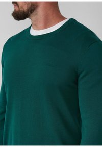 Dunkelgrüner Rundhalsausschnitt-Pullover aus weichem Stoff mit strukturierter Oberfläche und einem kleinen Logo-Detail auf der Vorderseite in der Nähe des Kragens.