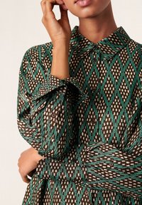 Camicia a fantasia marrone e teal con colletto, caratterizzata da forme di diamante e accenti a pois. Polsini abbottonati con tessuto testurizzato.