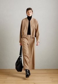 Beige bomberjacka över en svart polotröja, kombinerad med vida beiga byxor. Svarta läderankelstövlar och en texturerad svart väska.