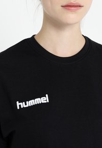 Young person wearing a black crewneck shirt with white "hummel" logo on chest, lips closed, on a plain white background.