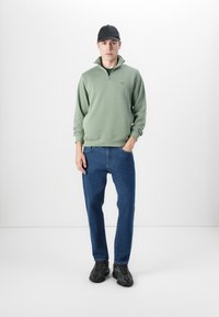 Groene hoodie met rits en hoge kraag, gecombineerd met blauwe spijkerbroek en zwarte sneakers, gedragen door een model tegen een egale achtergrond.