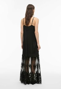 Femme aux longs cheveux bruns portant une robe noire sans manches avec un voile en dentelle florale transparente de mi-cuisse jusqu'au sol, vue de dos.