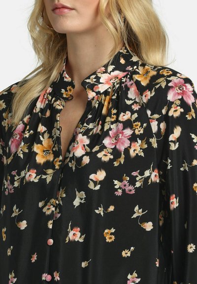 Femme blonde portant un chemisier noir à imprimés floraux roses, jaunes et oranges, avec un col haut froncé.