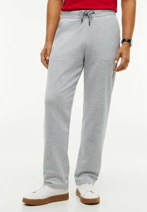 Personne portant un pantalon de jogging gris clair avec une taille à cordon, des baskets blanches à semelles marron, un t-shirt rouge, et une montre-bracelet.