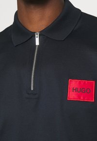 Gros plan sur un polo noir à zip avec un patch logo carré rouge portant l'inscription "HUGO" sur la poitrine.