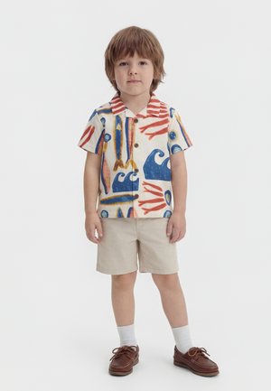 Niño pequeño de pie vestido con camisa de manga corta estampada con olas y peces, pantalones cortos beige, calcetines blancos y zapatos marrones sobre fondo blanco.