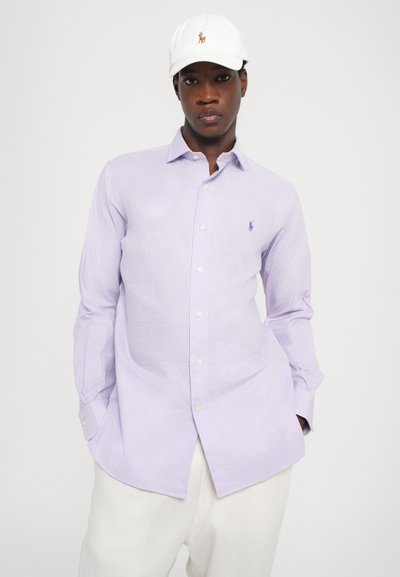 Cămașă cu carouri de un violet deschis, cu mâneci lungi, guler cu butoane și broderie logo pe pieptul stâng. Asortată cu pantaloni albi și o șapcă.