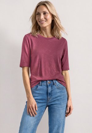 Femme blonde portant un t-shirt à manches courtes rose foncé et un jean bleu, debout avec une main dans la poche, souriant et regardant sur le côté.