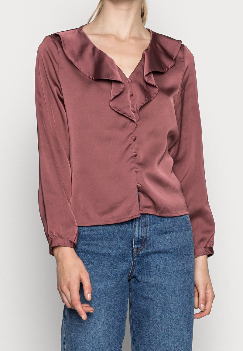 Femme portant un chemisier en satin mauve avec un col en V à volants et des boutons, associé à un jean en denim bleu taille haute, posant devant un fond uni.