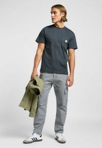 T-shirt grigio scuro con tasca frontale, abbinato a pantaloni cargo grigi chiari. Il modello tiene in mano una giacca verde oliva e indossa sneaker nere e bianche.
