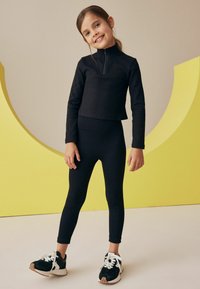 Maglia nera a costine con maniche lunghe e mezze zip, abbinata a leggings aderenti neri. Il bambino indossa scarpe da ginnastica bianche e nere. Sfondo giallo.