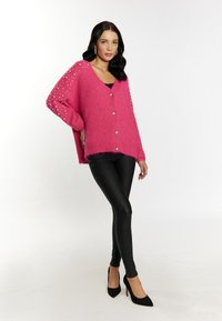 Cardigan oversize fucsia con dettagli in perle sulle maniche, dotato di scollo a V e chiusura con bottoni, abbinato a leggings neri e tacchi.