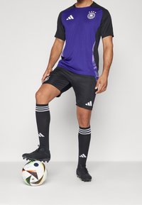 Lila och svart fotbollströja med korta ärmar, prydd med ränder och laglogotyp, kombinerad med svarta shorts och knähöga randiga strumpor.