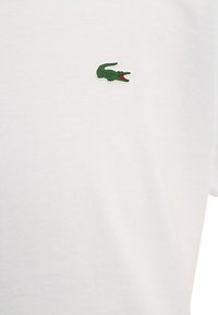 Vit tyg med en liten grön krokodil-logotyp som har röd färg inuti sin öppna mun, placerad på övre bröstområdet.