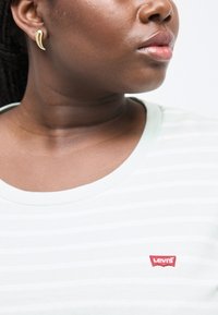 Κοντινό πλάνο ενός ατόμου που φορά χρυσά κυρτά σκουλαρίκια και ανοιχτόχρωμο ριγέ πουκάμισο Levi's, κοιτάζοντας προς τα δεξιά.