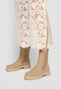 Bottines en suède beige avec des panneaux latéraux en élasthanne, des semelles en caoutchouc et un design chunky, associées à une robe en dentelle à motifs floraux.