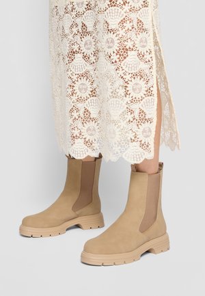Bottines en suède beige avec des panneaux latéraux en élasthanne, des semelles en caoutchouc et un design chunky, associées à une robe en dentelle à motifs floraux.
