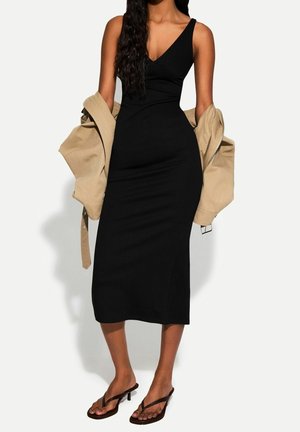 Femme portant une robe midi noire sans manches, tenant une veste beige sur ses bras, associée à des sandales à talons noires.