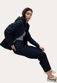 Černá prošívaná péřová bunda s kapucí, pod ní pruhované tričko, k tomu tmavé džíny a béžové tenisky s černými akcenty.