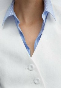 Gilet bianco con due bottoni rotondi sopra una camicia a righe azzurre, caratterizzato da una scollatura a V profonda e un design del colletto morbido.