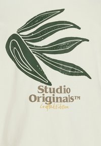 Kremna majica s velikim zelenim grafičnim motivom lista in napisom "Studio Originals™ Crafted Edition" v rjavi in rumeni barvi. Mehka tekstura.