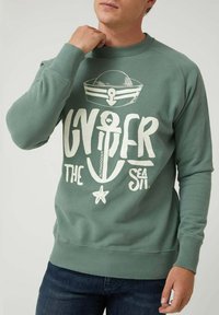 Hombre con sudadera verde con diseño náutico blanco, ancla, gorra de marinero y texto "Sobre el Mar."