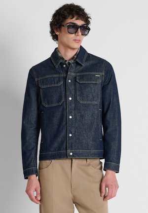 Uomo che indossa una giacca di jeans blu scuro con bottoni a pressione, pantaloni beige, camicia a quadri e grandi occhiali da sole neri, in piedi su uno sfondo chiaro.