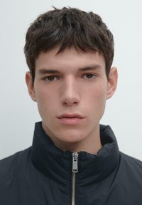 Czarna kurtka na zamek błyskawiczny z wysokim kołnierzem, wyposażona w metalowy zamek. Modelka ma krótkie, proste brązowe włosy i neutralny wyraz twarzy.