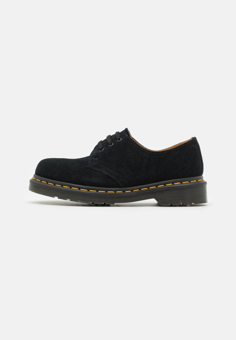 Chaussure basse en daim noir à lacets avec semelle épaisse et couture jaune le long du bord, sur un fond clair uni.