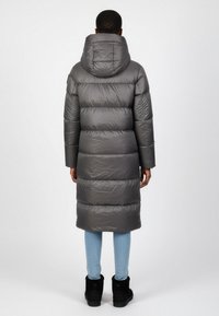 Lang grå jakke i puffer stil med hætte, med en quiltet design, blank overflade og længde til midten af læggen. Matcher med lyseblå jeans og sorte støvler.