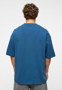 Overgrote t-shirt met korte mouwen in tealblauw, gemaakt van zachte stof met een ronde ronde hals en zichtbare stiksels, gedragen met grijze denimshorts.