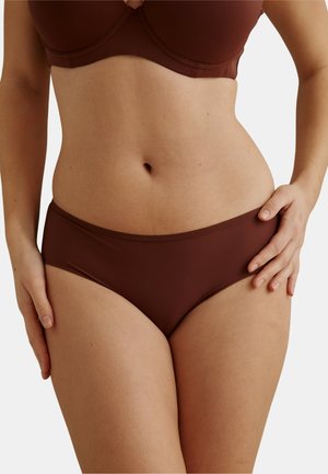Femme portant un soutien-gorge brun foncé et une culotte assortie, debout devant un fond clair uni, une main posée sur la hanche.