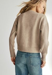 Pulover tricotat bej cu o croială lejeră, cu manșete și tiv ondulate. Asortat cu blugi de culoare albastră de tip high-waisted, uzati, care au buzunare la spate.