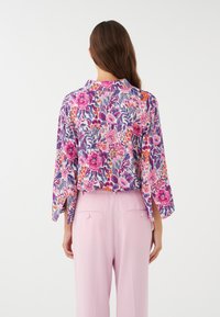 Blouse florale en violet, rose et orange avec des manches trois-quarts, arborant un motif distinct. Assortie à un pantalon rose clair.
