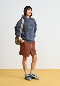 Felpa grigia con cappuccio e logo, gonna marrone al ginocchio con zip frontale, sneakers grigie e nere, e calzini alla caviglia color crema.